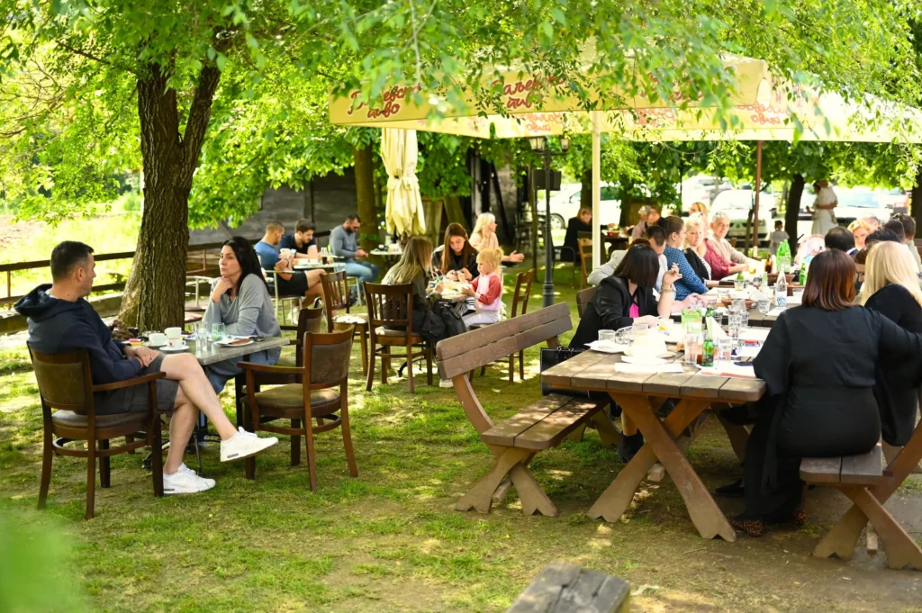Lepo vreme, zelenilo, priroda i ljudi koji sede u hladu ispod suncobrana i drveća.