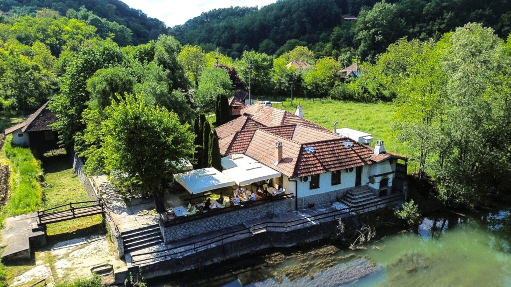 Slika iz vazduha prikazan restoran, lepo vreme i reka pored restorana ljudi sede u hladu
